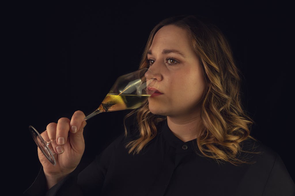 A serene woman in black thoughtfully sips white wine against a dark background.