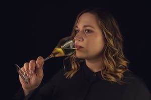 A serene woman in black thoughtfully sips white wine against a dark background.