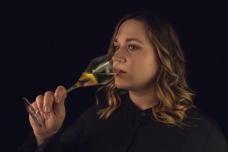 A serene woman in black thoughtfully sips white wine against a dark background.