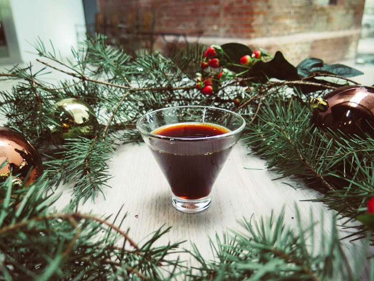 A glass of dark holiday drink encircled by Christmas decorations and pine greenery on a table.
